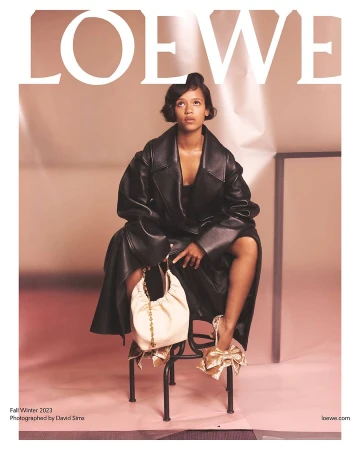 Fashion editorial featuring a woman in a black leather coat, sitting on a metal chair, holding a cream-colored bag in a studio setting.