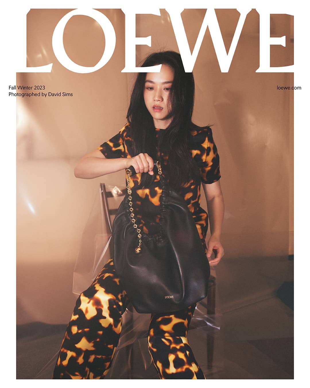 Fashion editorial featuring a model holding a black Loewe purse, posing indoors in a patterned suit, with the brand logo visible.