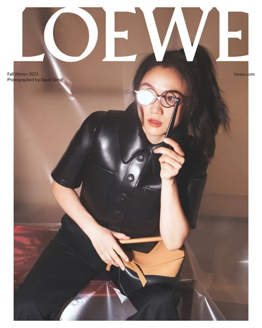 Editorial fashion shot featuring an Asian woman in a black leather top and pants, holding a tan handbag and wearing glasses, against a neutral backdrop.