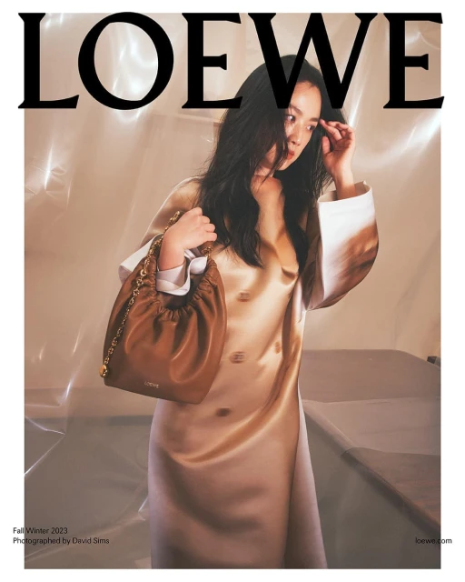 Fashion editorial shot of an Asian female model in a champagne dress holding a brown leather purse by Loewe in a bright studio.