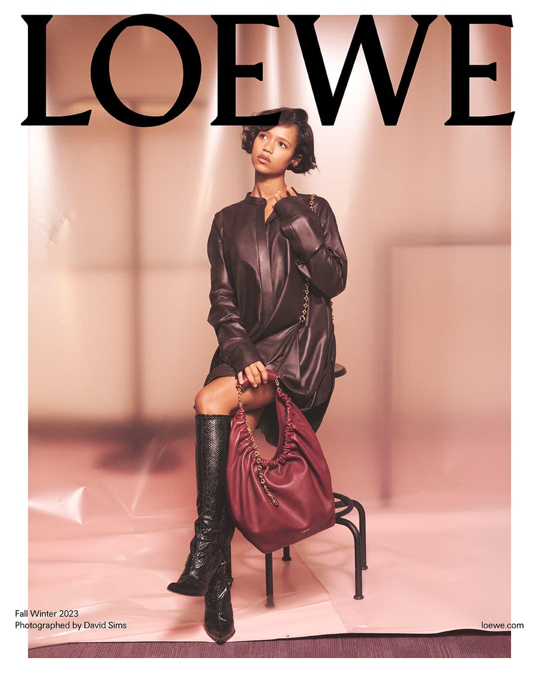 A woman models Loewe Fall/Winter 2023 collection, seated with a burgundy handbag and wearing knee-high snakeskin boots.
