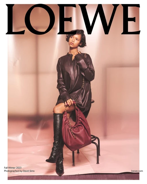A woman models Loewe Fall/Winter 2023 collection, seated with a burgundy handbag and wearing knee-high snakeskin boots.