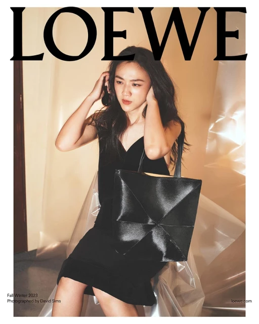 Fashion editorial shot of a young woman in a black dress, posing with a black leather tote bag, featuring the LOEWE logo.
