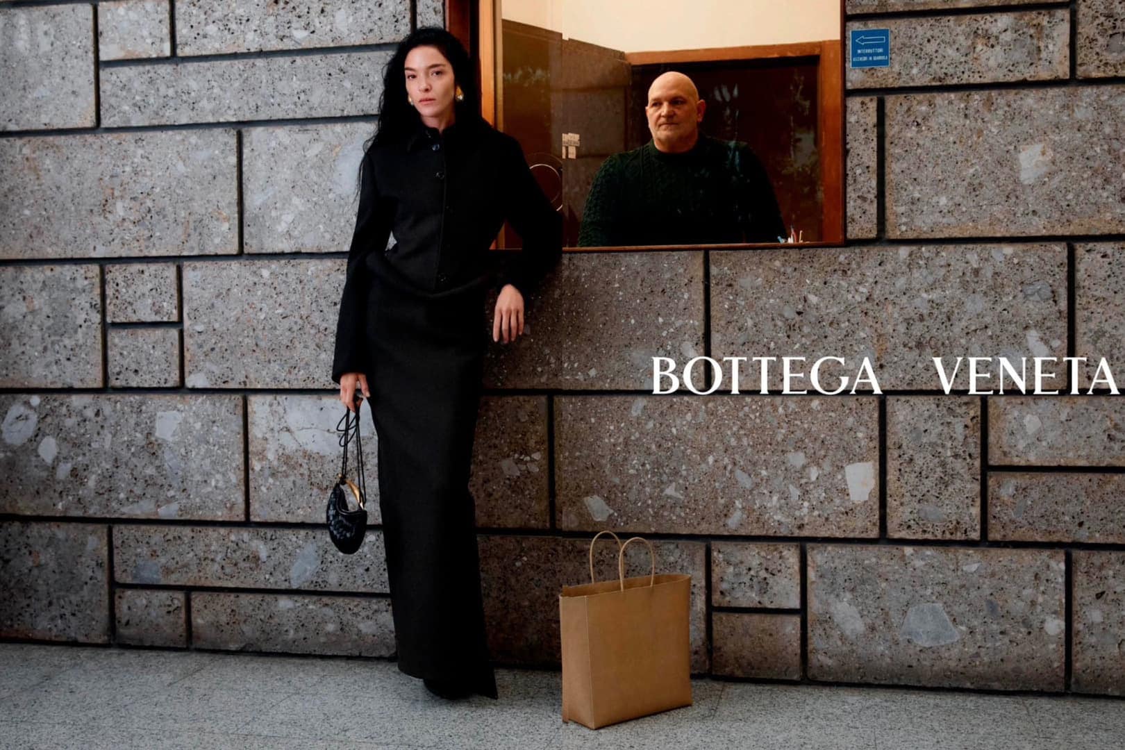 Fashion editorial shot of a woman in black leaning against a brick wall, with an older man visible through an opening.