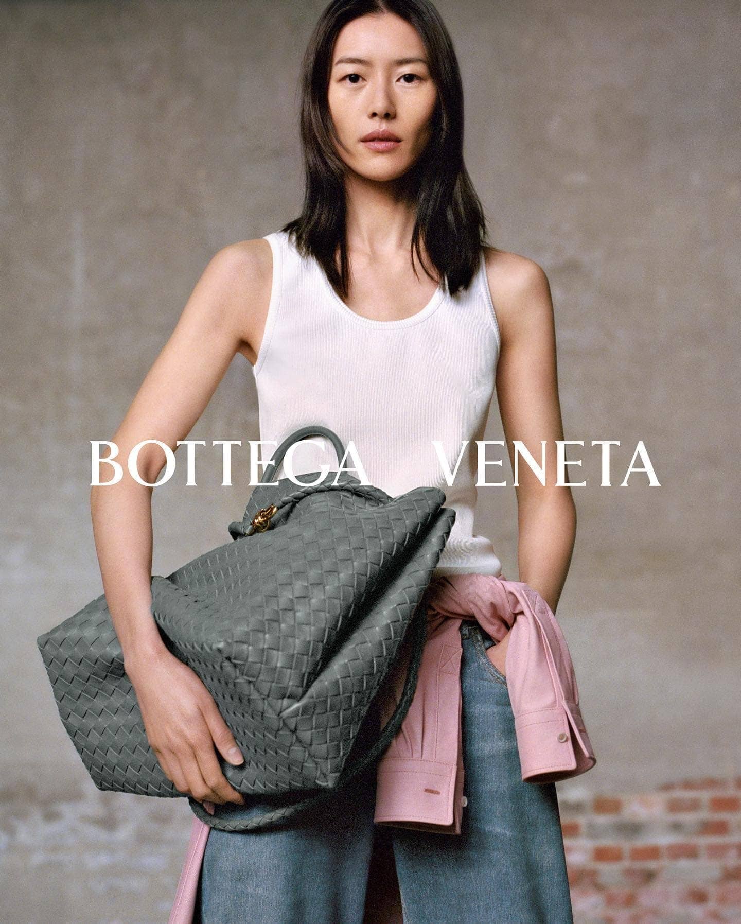 Asian woman posing with a woven gray leather tote bag, wearing a white top and jeans, with a pink shirt draped over her arm.