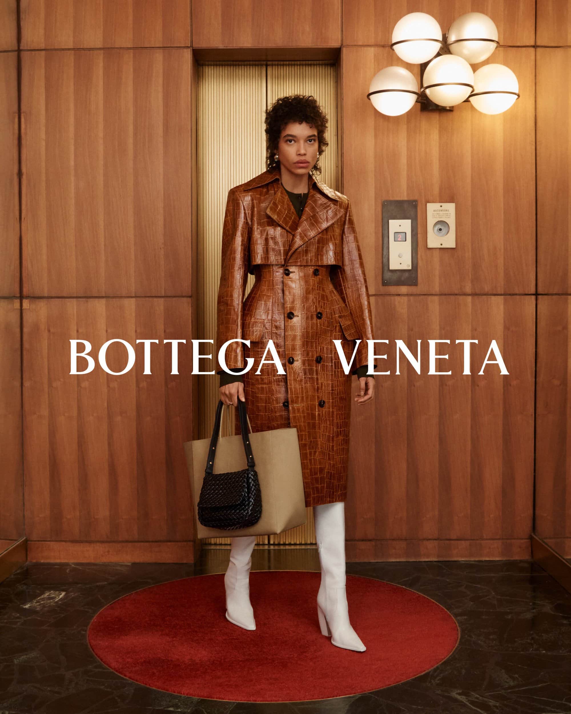 Woman in a brown trench coat and white boots holding a tote bag, standing in front of an elevator with wood paneling in a fashion shot.