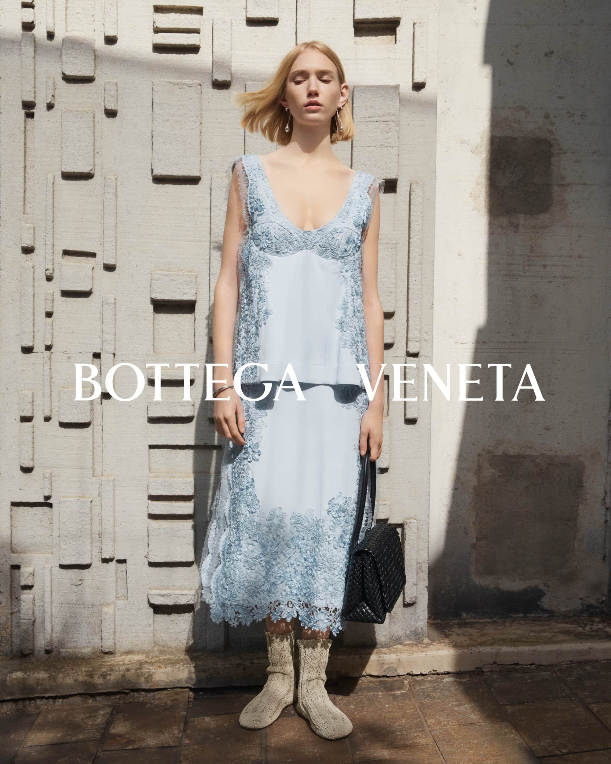 A fair-skinned fashion model poses in a light blue lace dress and wool ankle boots with a black woven handbag in a studio shot.