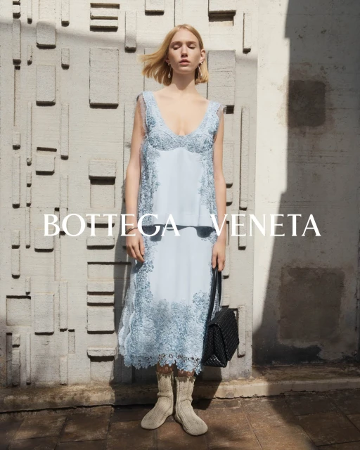 A fair-skinned fashion model poses in a light blue lace dress and wool ankle boots with a black woven handbag in a studio shot.