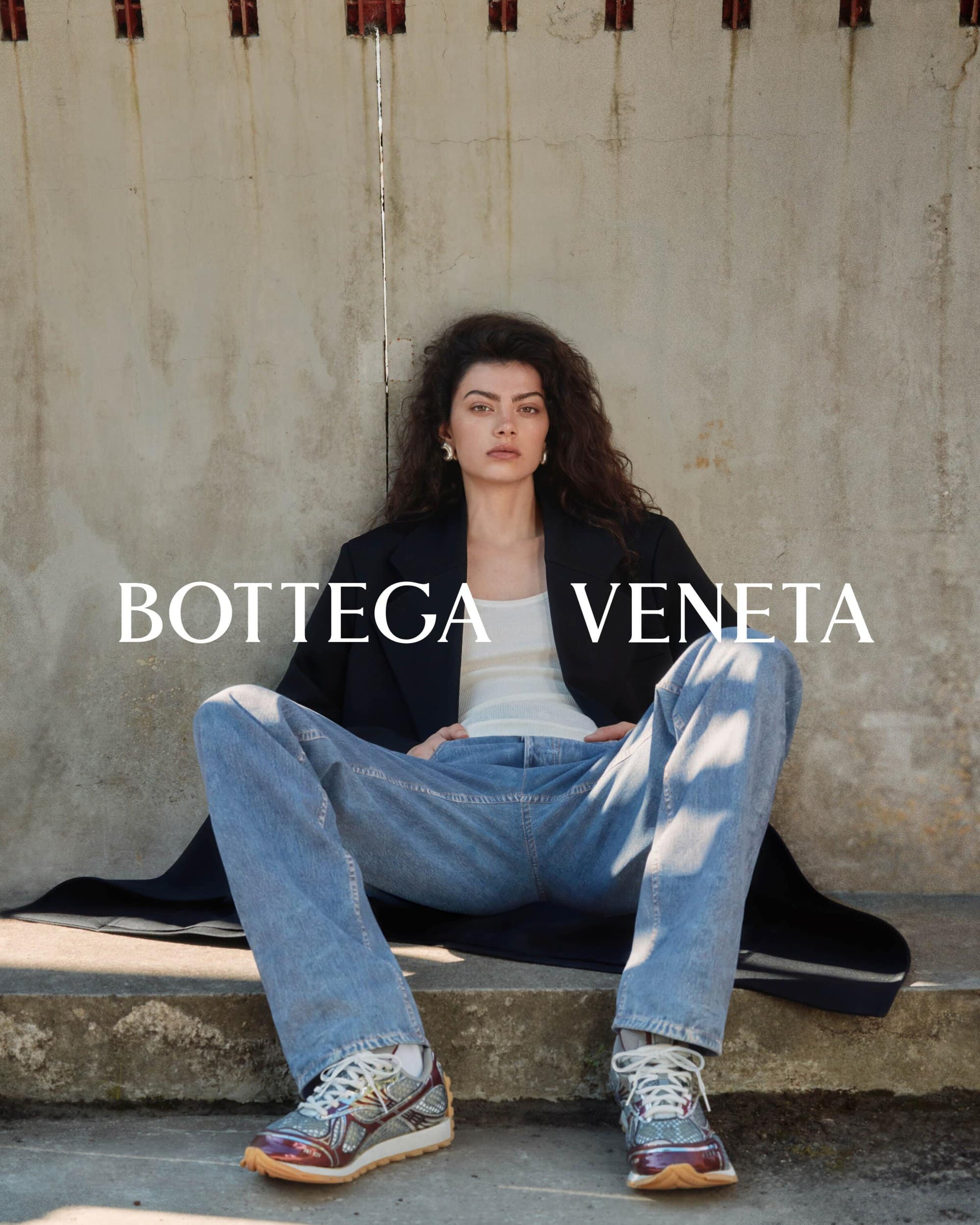 A fashionable woman in jeans and a blazer poses on concrete stairs with Bottega Veneta text overlay, showcasing casual elegance.