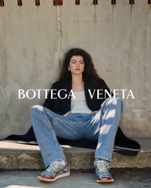 A fashionable woman in jeans and a blazer poses on concrete stairs with Bottega Veneta text overlay, showcasing casual elegance.