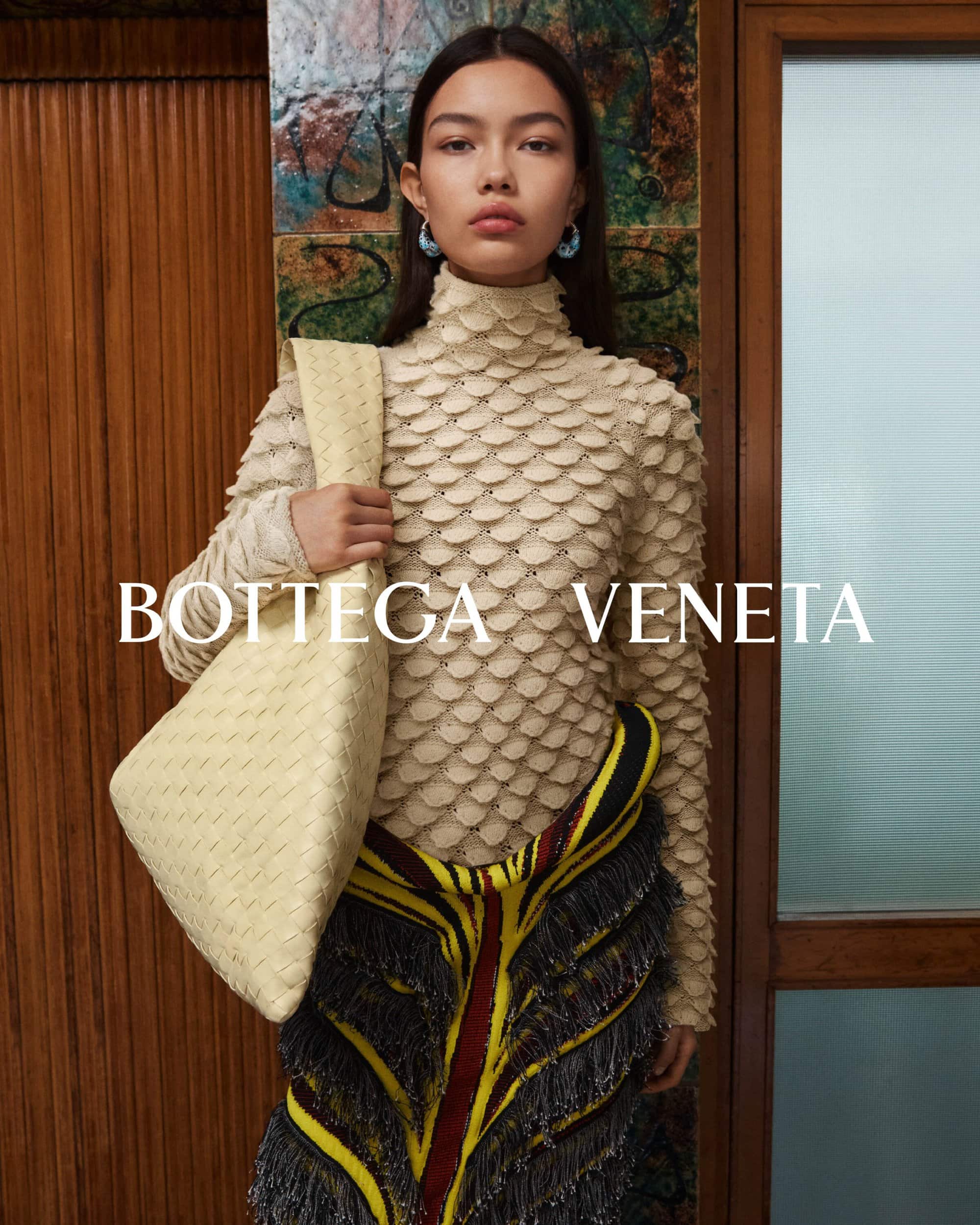 Editorial fashion shot features a woman wearing textured top and decorative skirt, holding a woven handbag in front of a wood-paneled wall.