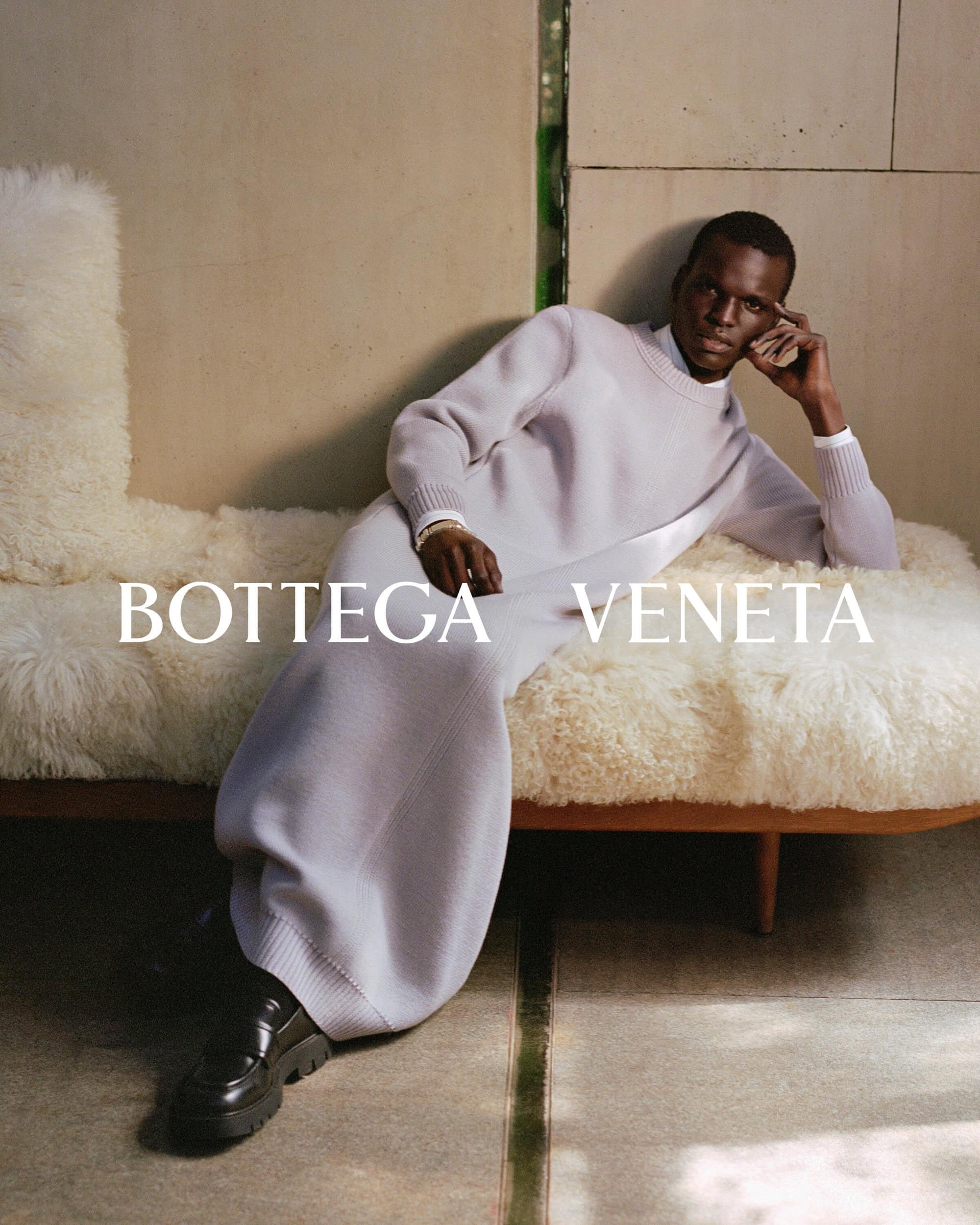 An adult Black male model reclines on a bench wearing a lavender sweater dress and black shoes, showcasing Bottega Veneta fashion.