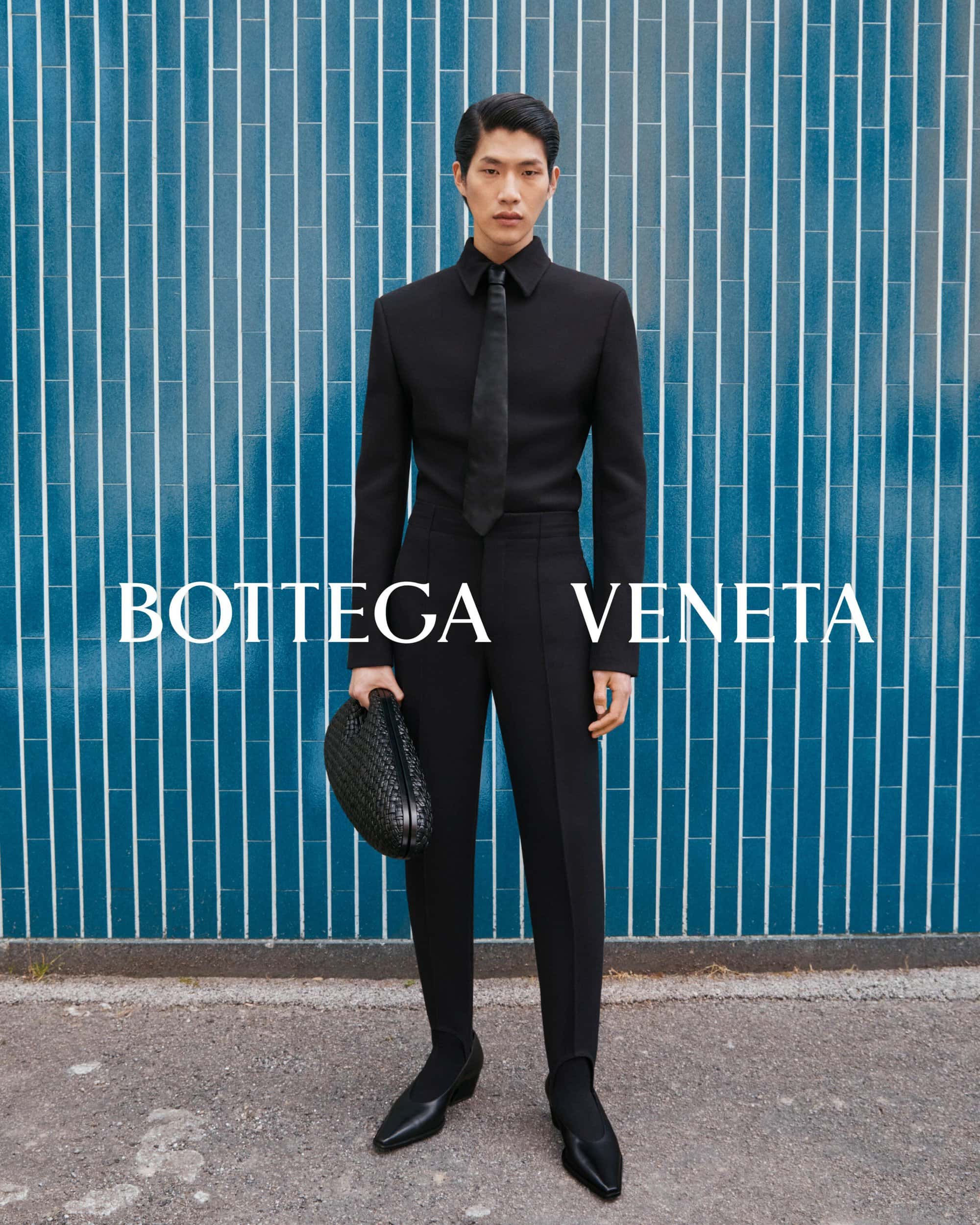 Asian man in a black suit posing against a blue tiled wall, holding a black woven bag, in a full shot for a Bottega Veneta ad.
