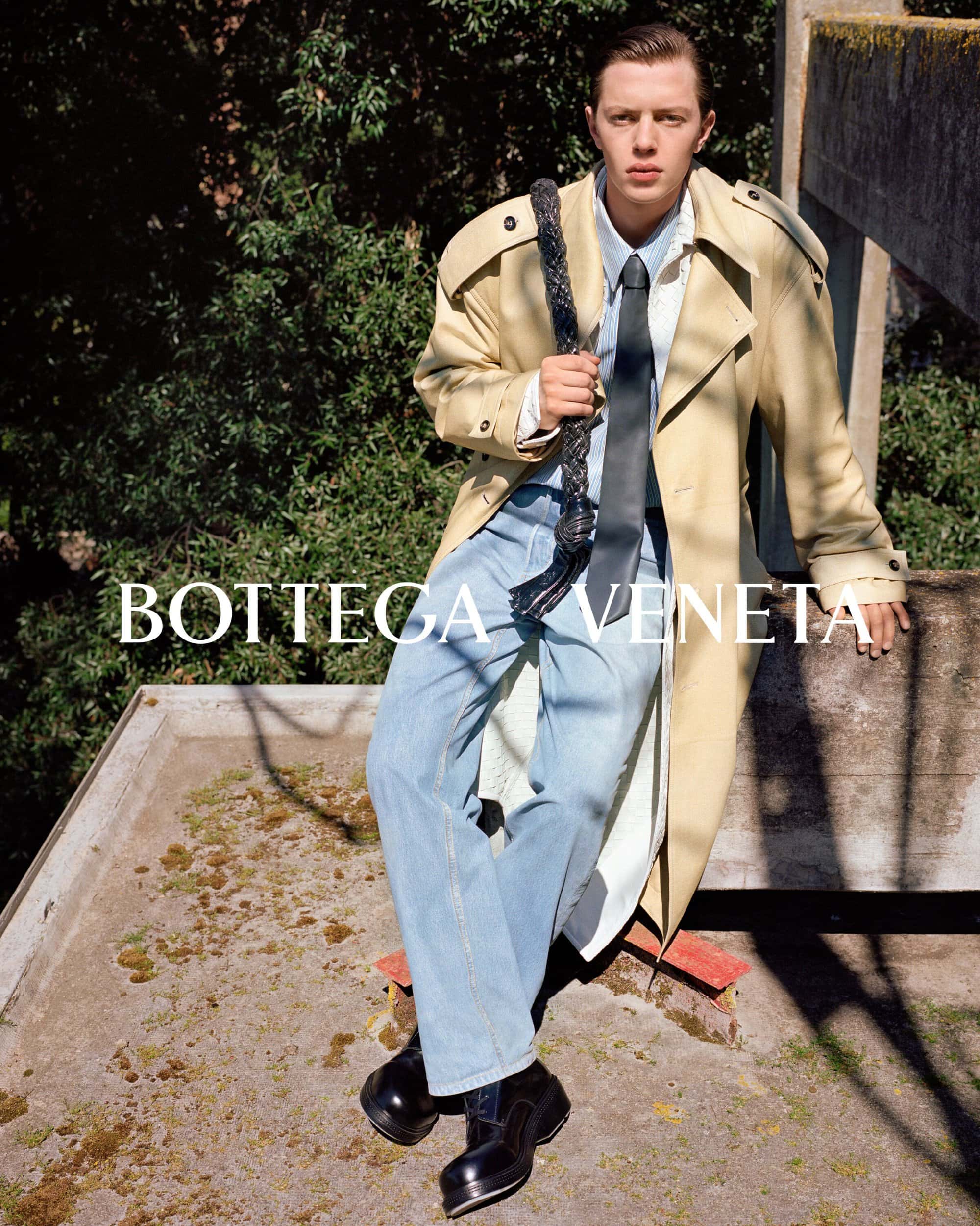 Fashion editorial shot of a young adult male in a trench coat, jeans, tie, and platform shoes, sitting on a concrete surface outdoors.