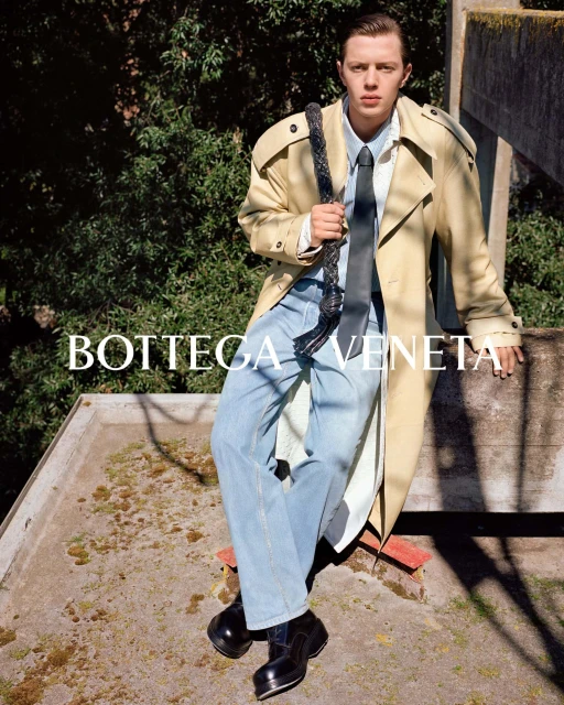 Fashion editorial shot of a young adult male in a trench coat, jeans, tie, and platform shoes, sitting on a concrete surface outdoors.