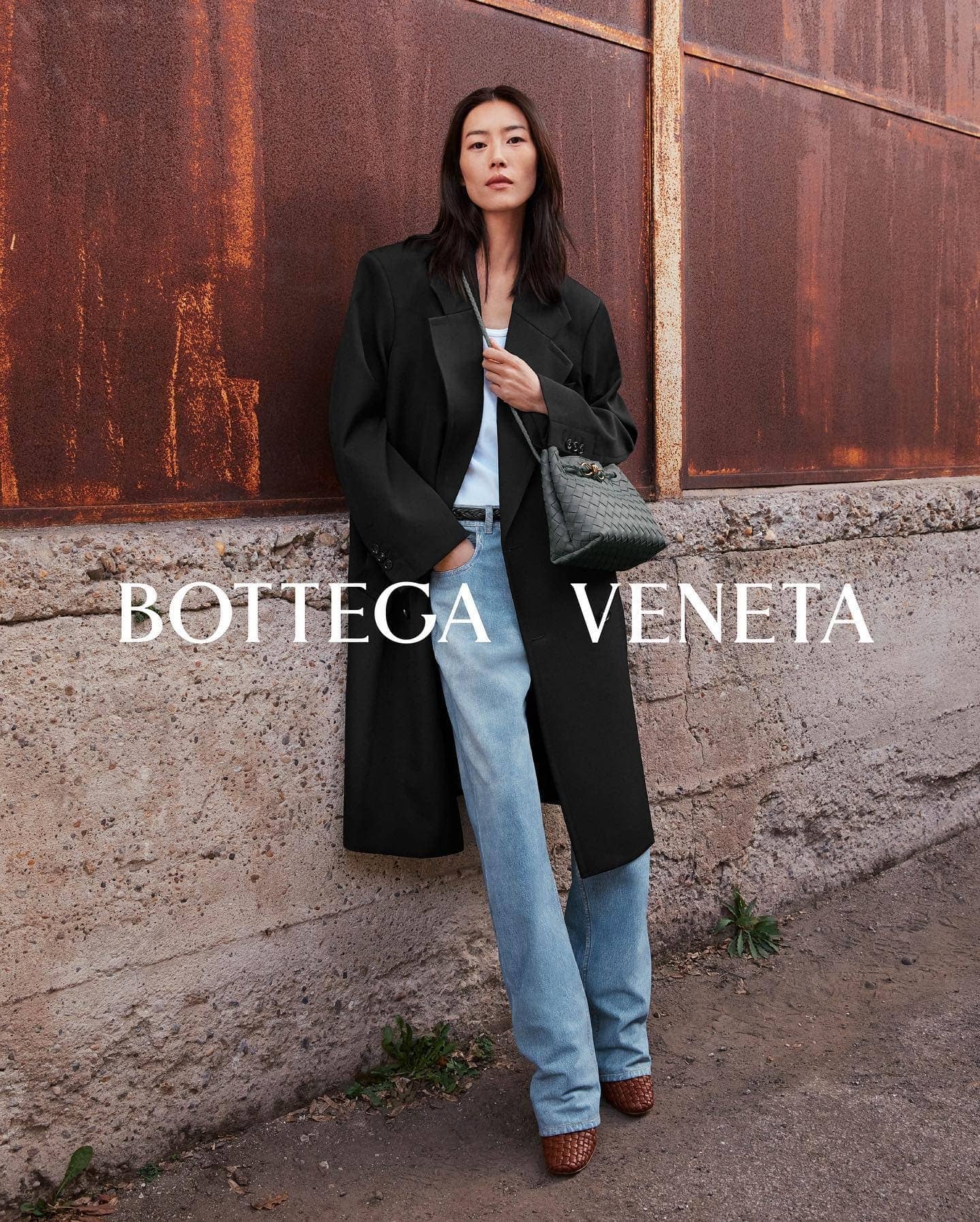 Asian woman poses in a black coat and blue jeans against a rusted metal wall, holding a gray Bottega Veneta purse on a city street.