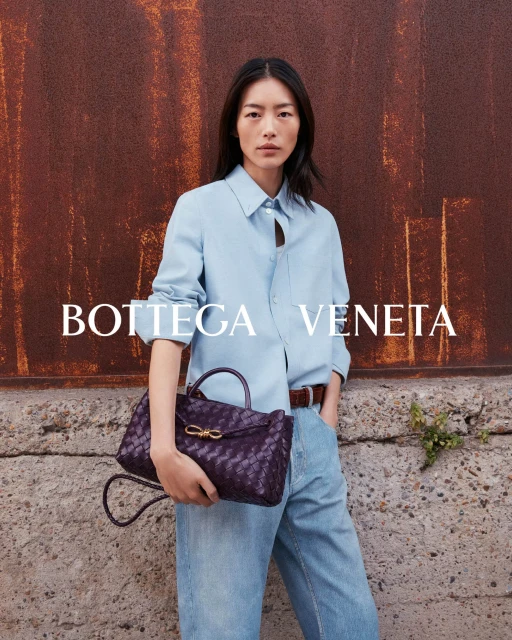 Woman wearing blue shirt and jeans holding Bottega Veneta handbag in front of rusted metal wall outdoors in commercial setting.