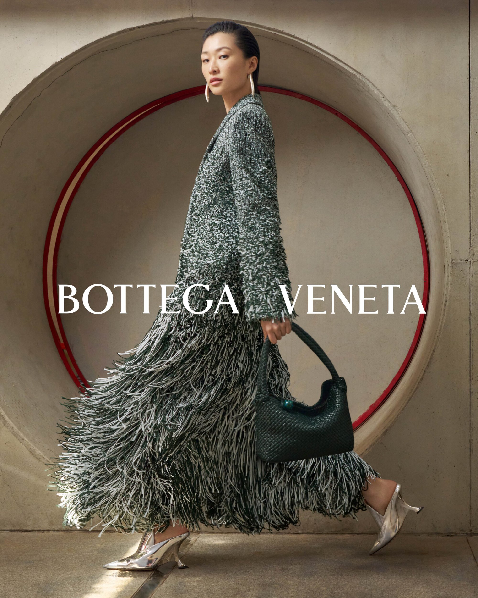 Fashionable Asian woman in a tweed coat and fringed skirt walks past a circular opening, carrying a woven bag and wearing heeled shoes.