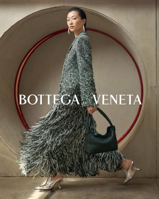 Fashionable Asian woman in a tweed coat and fringed skirt walks past a circular opening, carrying a woven bag and wearing heeled shoes.
