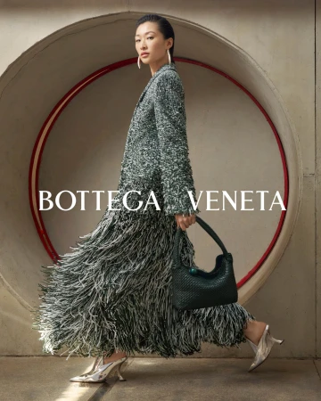Fashionable Asian woman in a tweed coat and fringed skirt walks past a circular opening, carrying a woven bag and wearing heeled shoes.