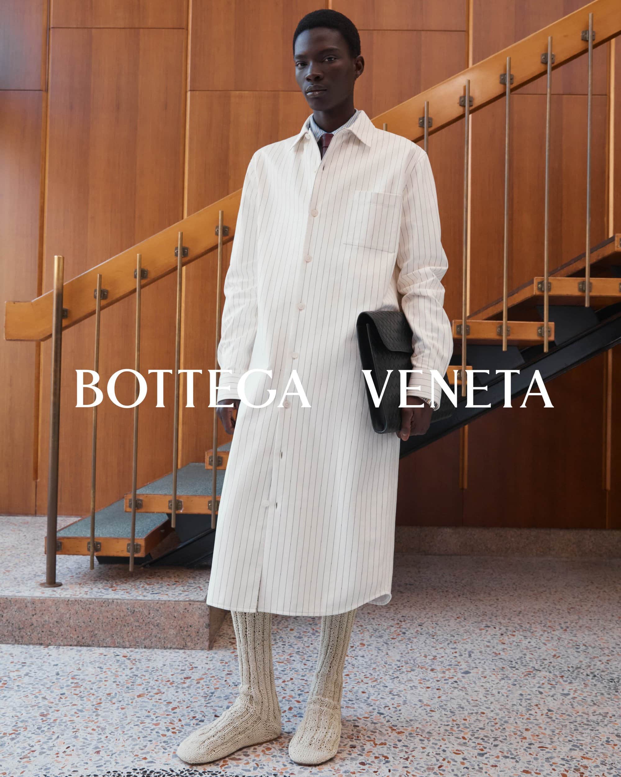 Model wearing a long white striped button-down coat and knit shoes standing in front of a wooden staircase for Bottega Veneta.