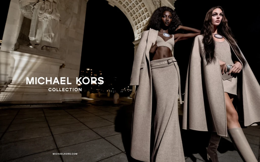 Two fashion models showcasing Michael Kors collection outdoor at night. The women wear tan outfits and pose near an archway.