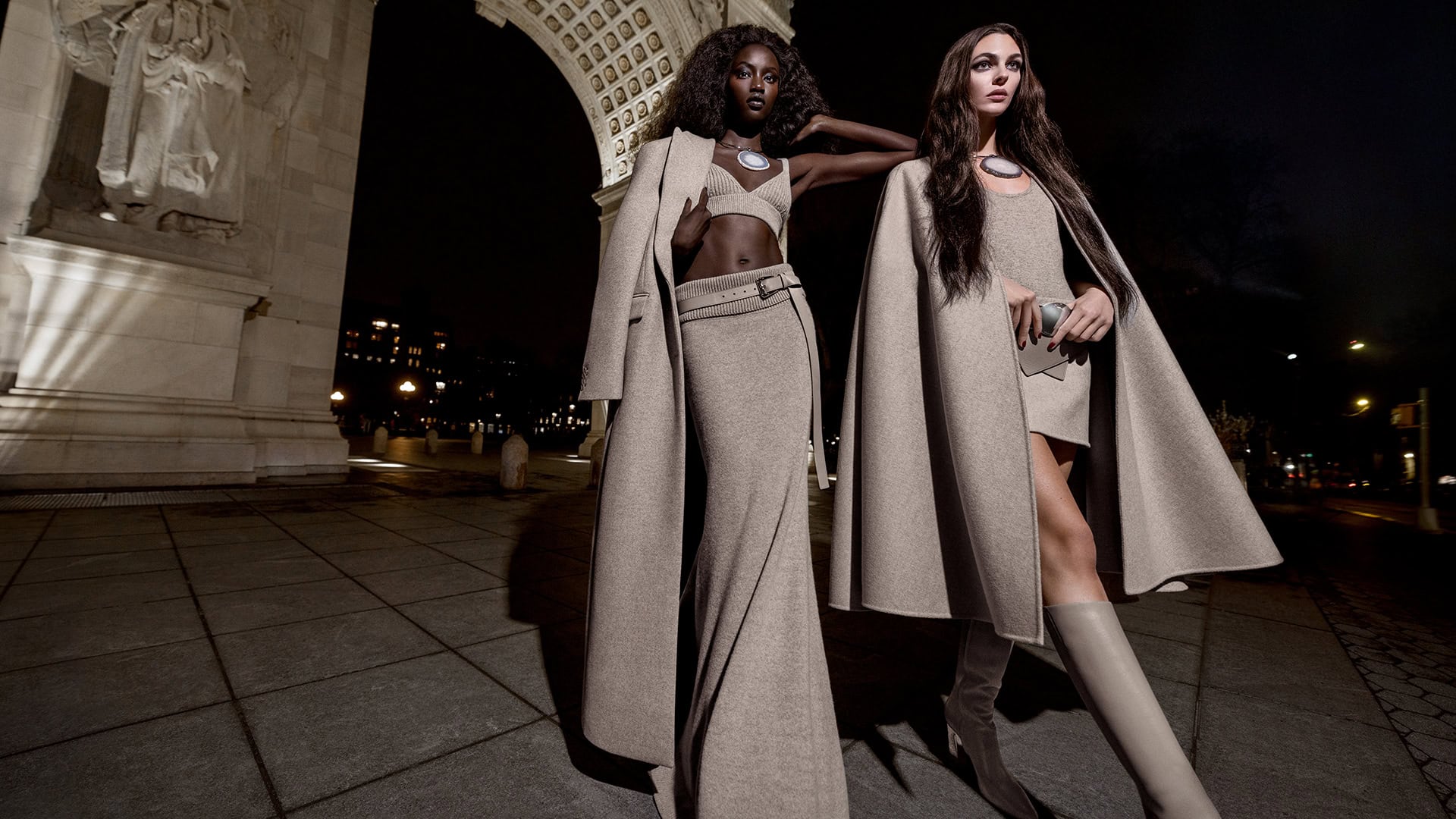 Two women model tan knitwear on a city sidewalk at night, showcasing matching outfits in front of an archway.