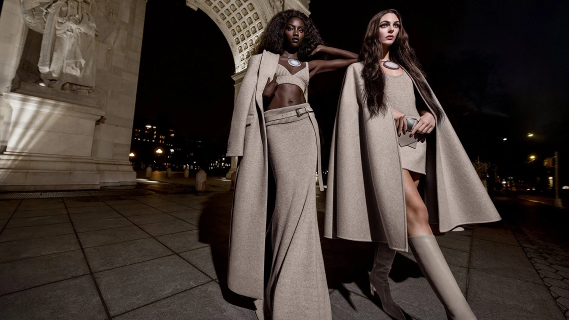 Two women model tan knitwear on a city sidewalk at night, showcasing matching outfits in front of an archway.