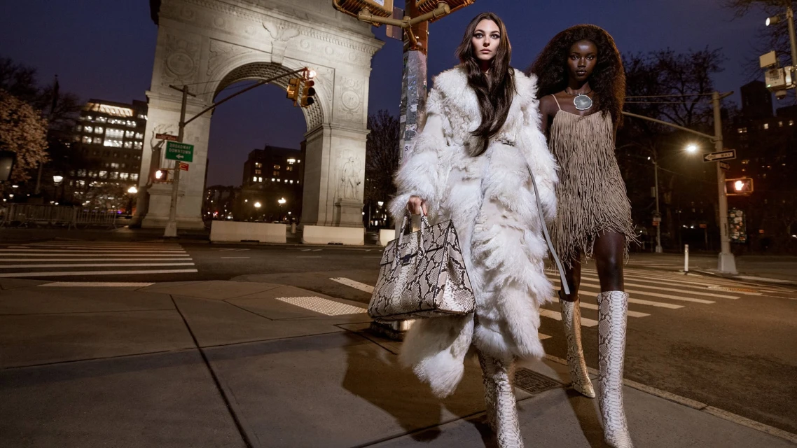 Two fashion models pose on a city street at night, with an archway, buildings, and streetlights in the background. One woman wears a fur coat.