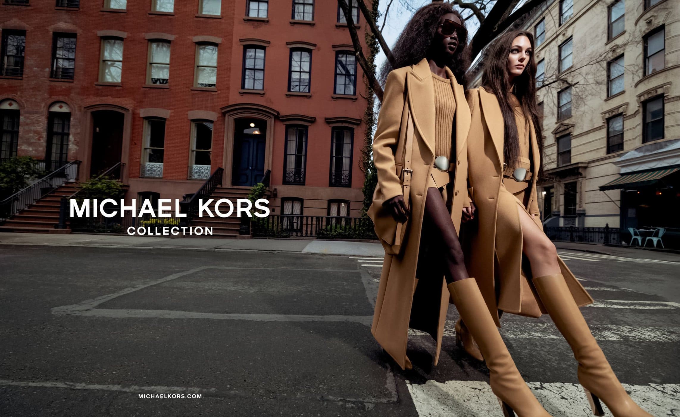 Two fashion models showcase tan trench coats from the Michael Kors collection, walking across a city street in front of brownstone buildings.
