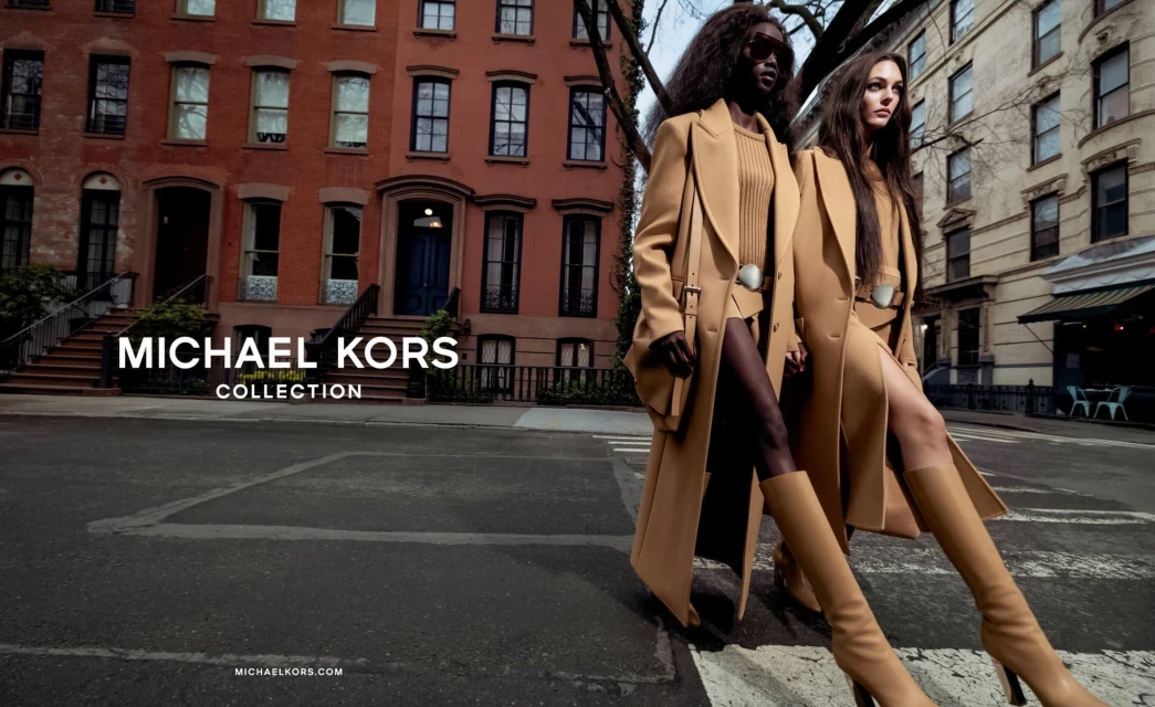 Two fashion models showcase tan trench coats from the Michael Kors collection, walking across a city street in front of brownstone buildings.