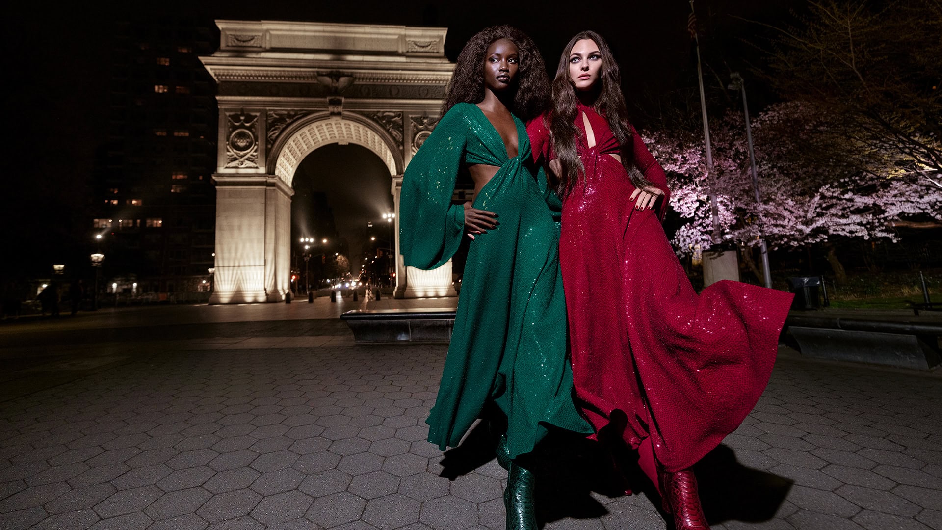 Two fashion models in evening gowns pose in front of the Washington Square Arch at night, showcasing a high-fashion aesthetic.