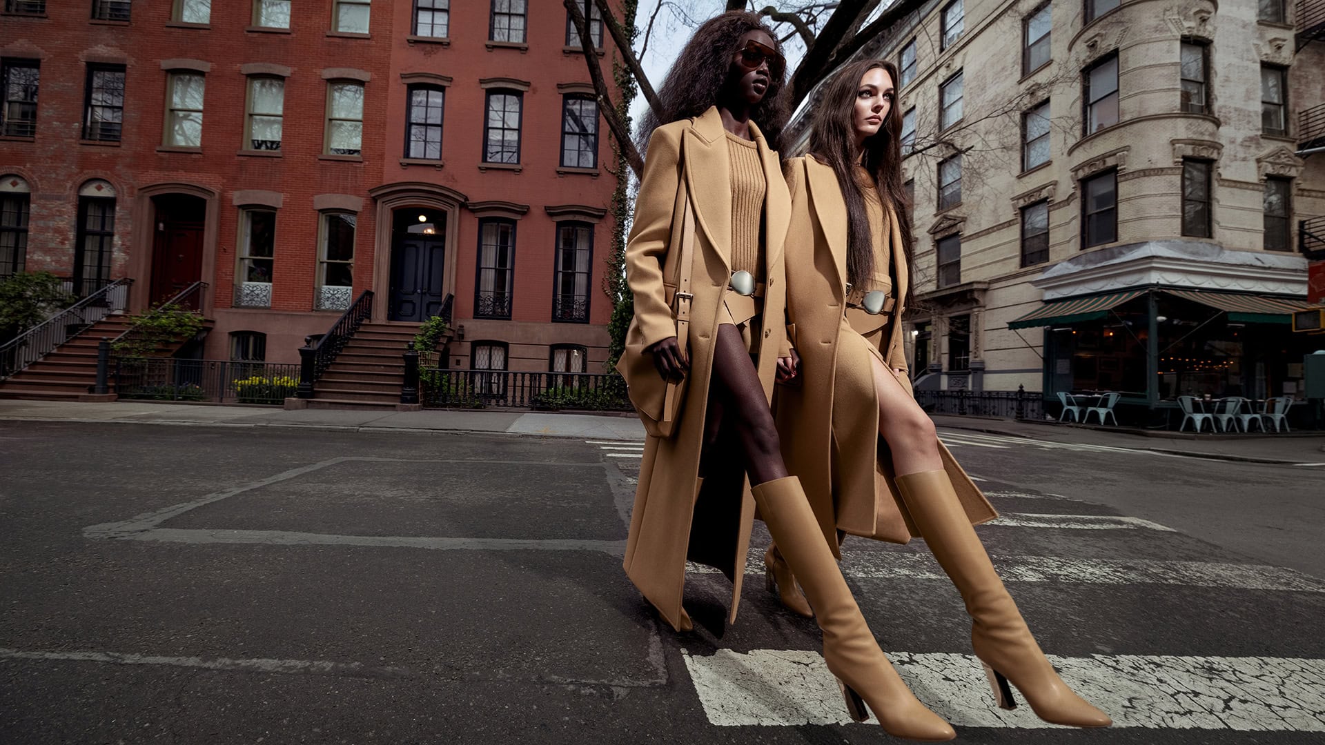 Two fashion models pose on a crosswalk in a city street wearing tan coats, boots, and matching outfits, with brick buildings in the background.