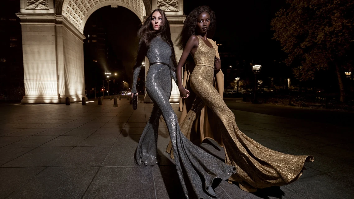Two fashion models wearing shimmering gowns pose in front of an archway at night, showcasing elegant evening wear and urban sophistication.