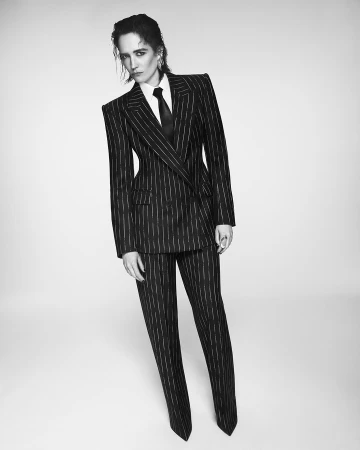 A woman with wet-look hair wears a sharp, pinstriped black suit with a white shirt and black tie against a minimalist studio background.