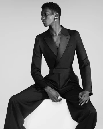 Momo Ndiaye seated wearing a tailored black double-breasted tuxedo jumpsuit with a cinched waist for Alexander McQueen.