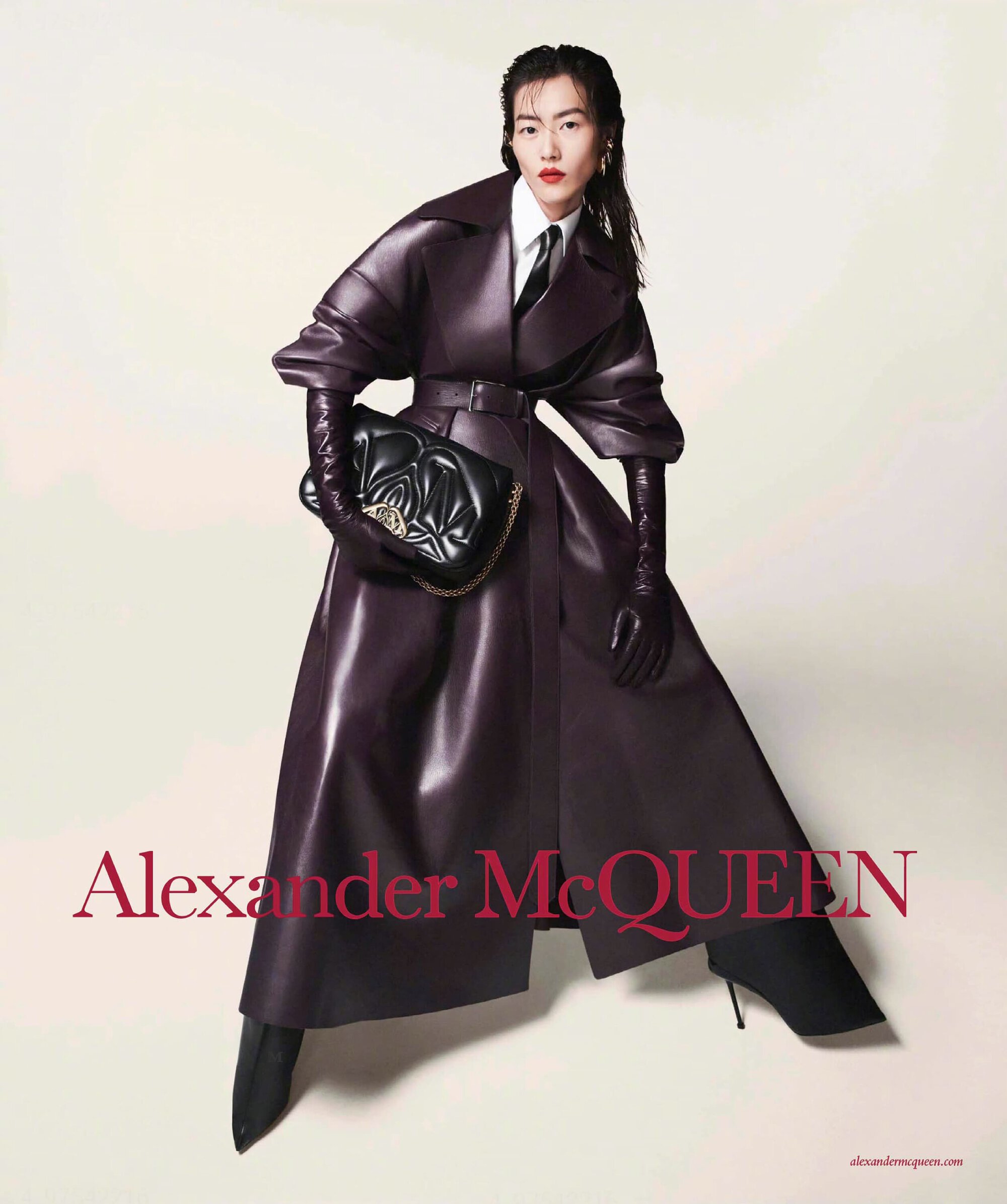 Fashionable woman in a dark purple leather coat with gloves, holding a black quilted shoulder bag in a studio with a light background.