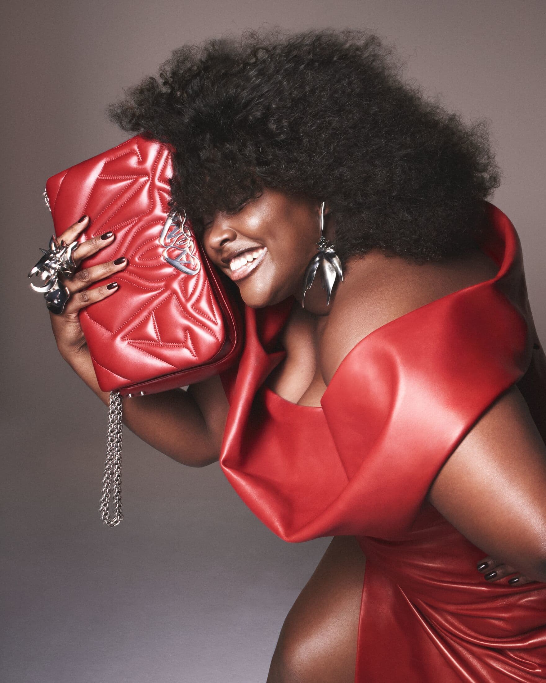 A smiling African American woman poses in a red dress, holding a red quilted purse with silver chain and accessories, against a grey background.