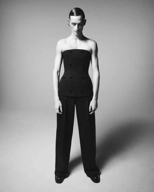 A slender adult stands in a studio. They are wearing a strapless black top, wide-leg black trousers, and patent-leather shoes. Minimalist style.