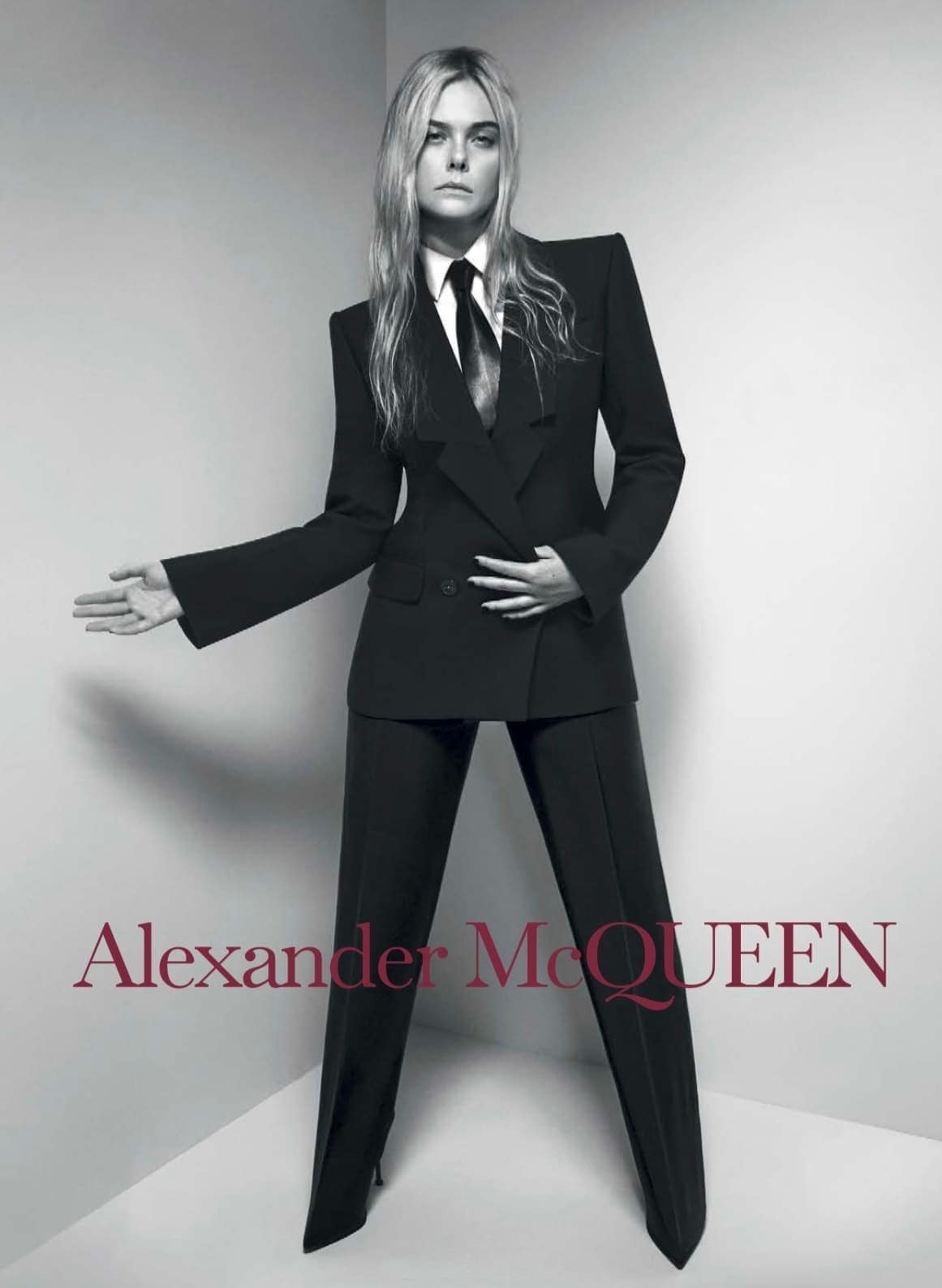 Elle Fanning poses in a black suit against a grey wall, wearing a tie. Shot in a photo studio for a fashion editorial, by Alexander McQueen.