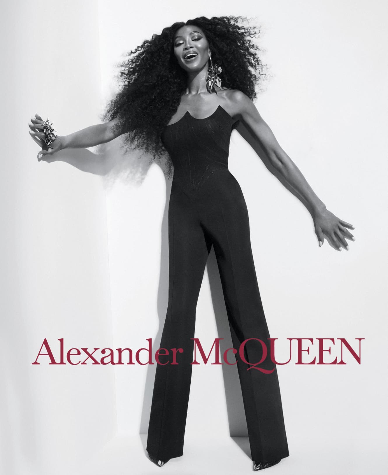 Fashion shot featuring an adult woman with dark hair wearing a black jumpsuit posing against a white background, promoting Alexander McQueen.