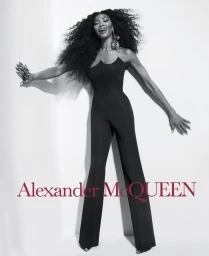 Naomi Campbell poses in a black strapless flared jumpsuit against a white studio wall with the red Alexander McQueen logo overlaid.