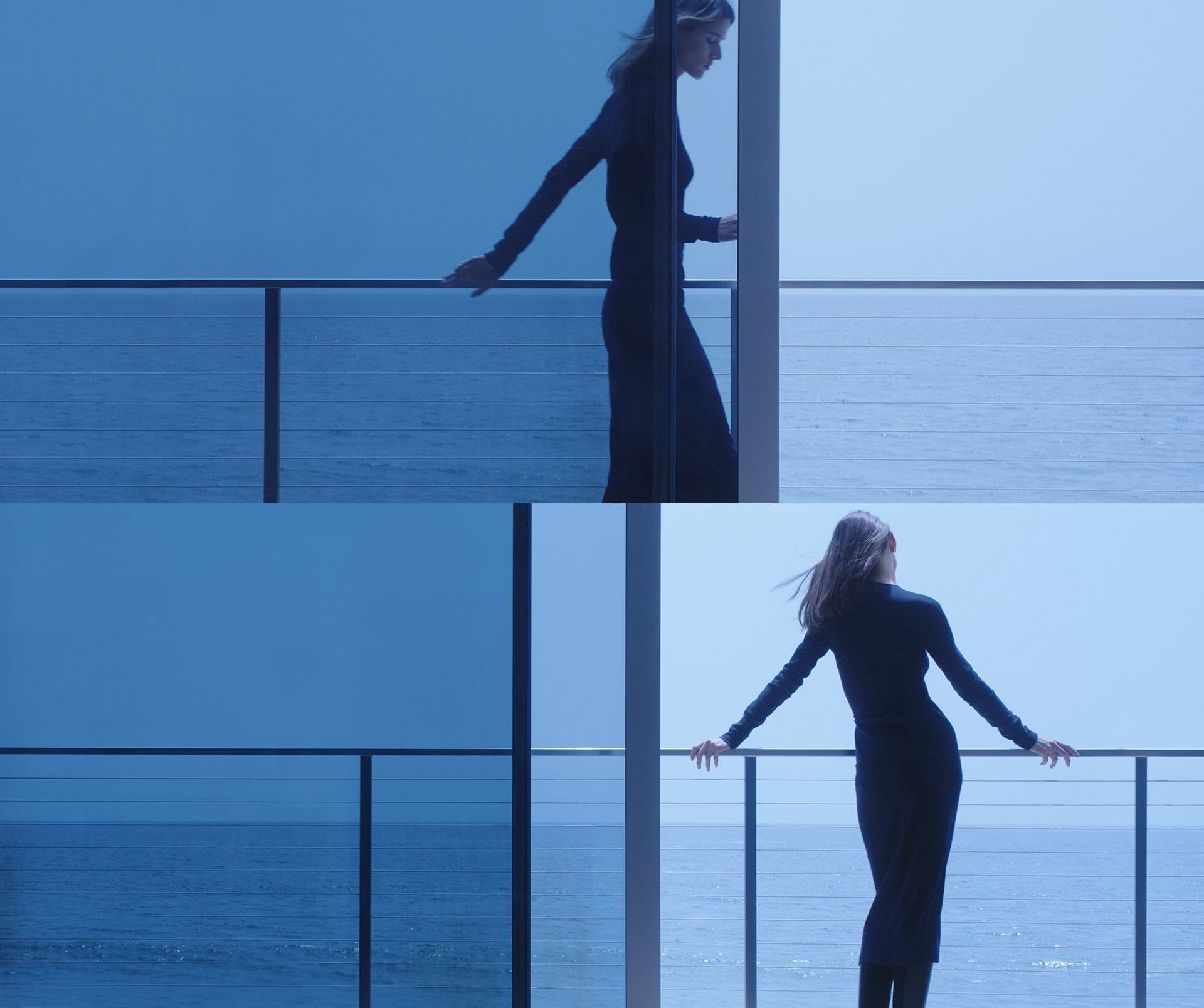 A woman in a long black dress stands on a balcony overlooking the ocean under a blue sky, gazing out at the water.