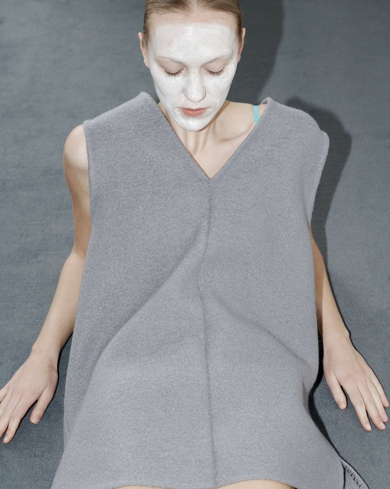 Woman with white face mask sits on a gray surface wearing a gray sleeveless shirt, creating a minimalist and neutral fashion image.