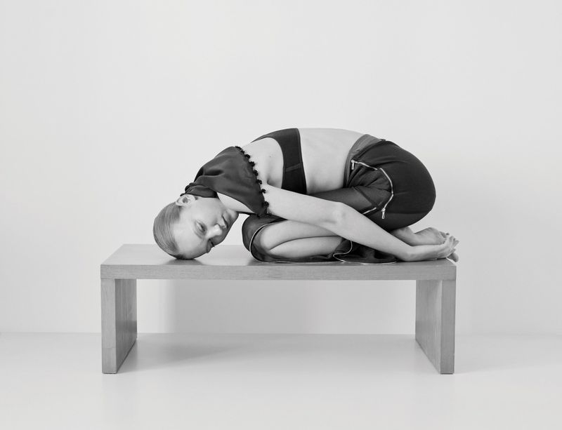 Monochrome studio shot of a Caucasian female model kneeling on a wooden table with her head down and arms wrapped around her legs.