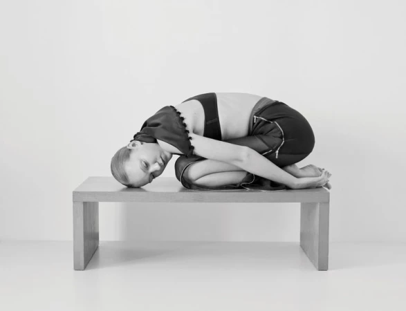 Monochrome studio shot of a Caucasian female model kneeling on a wooden table with her head down and arms wrapped around her legs.