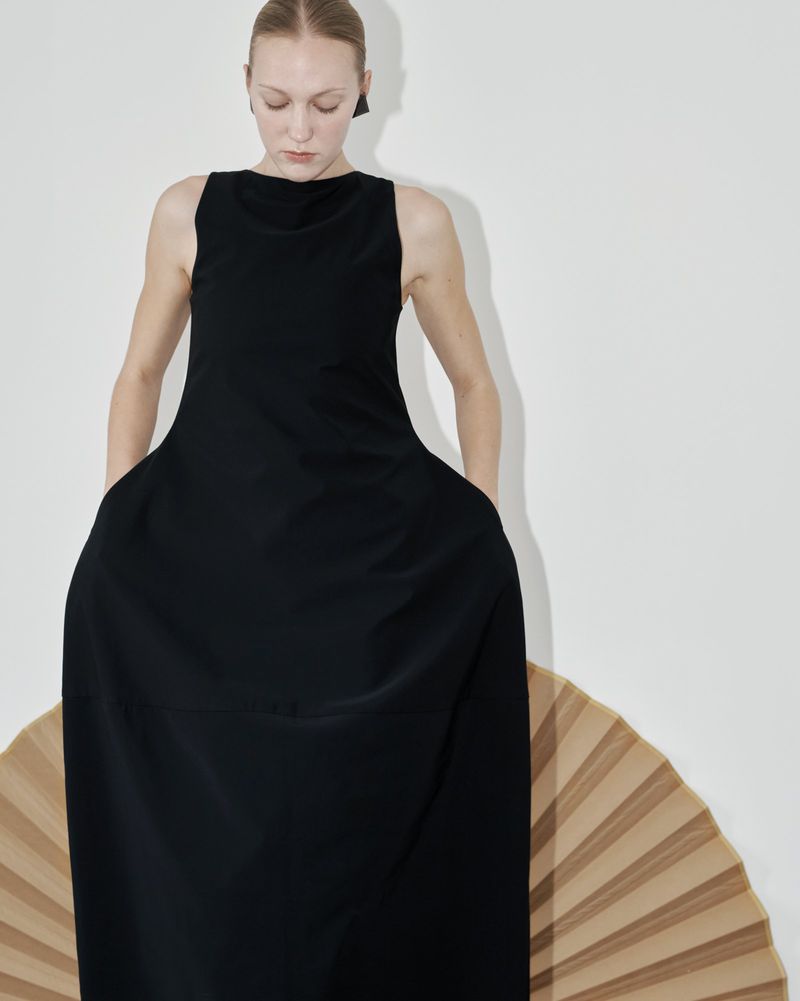 A fair-skinned woman wears a black sleeveless dress with a wide skirt, standing in front of a tan folding fan against a white wall.
