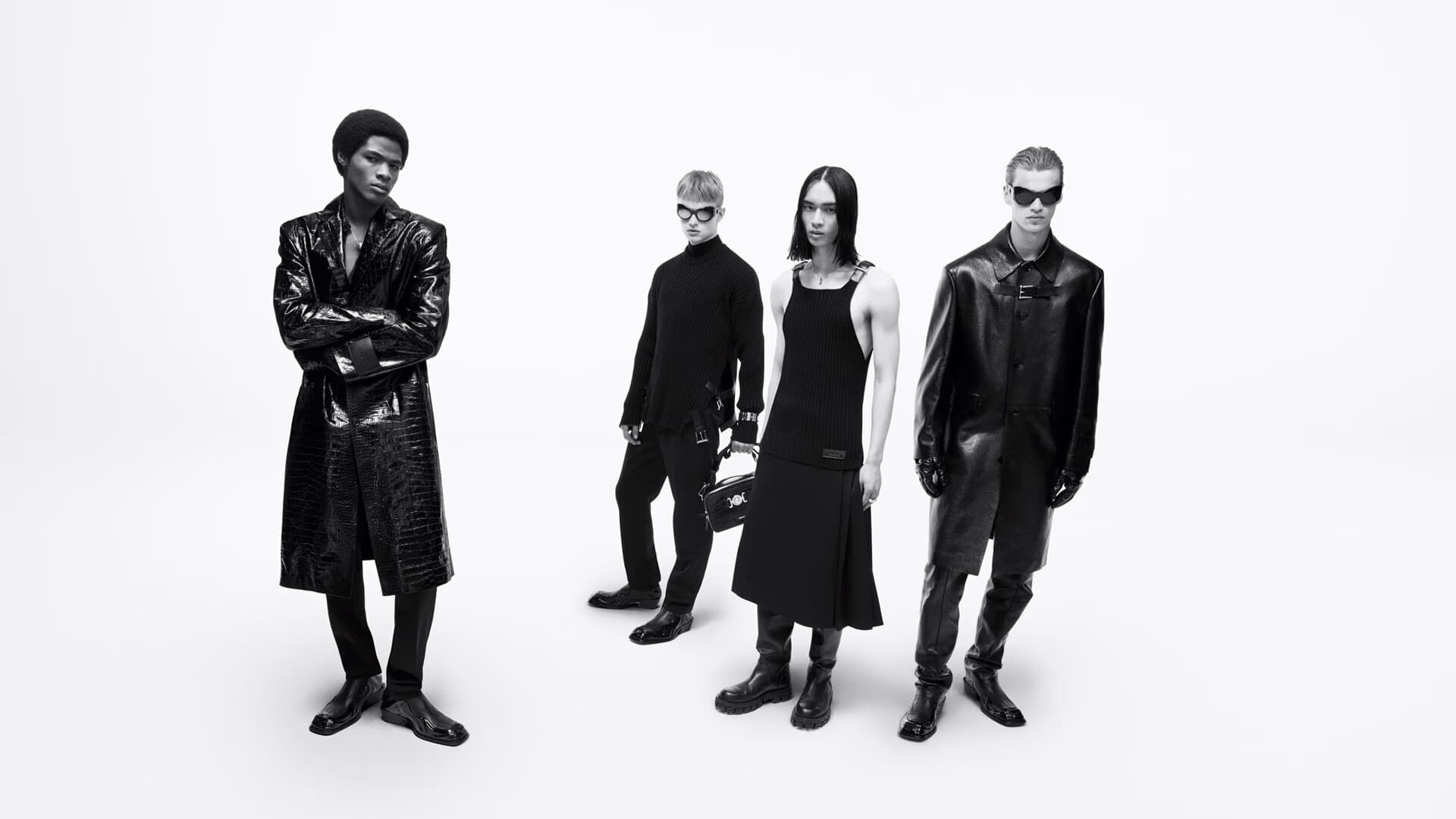 Four fashion models in an indoor studio setting wearing dark, futuristic clothing, including long coats, dresses, pants, and sunglasses.