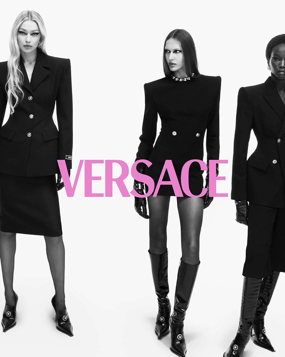 Three fashion models in black power-dressing suits, accessorized with heels and gloves, stand in a photo studio with the Versace logo.
