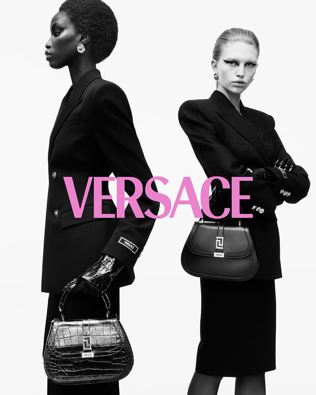 Two fashion models in black suits showcase Versace handbags against a bright background in a studio fashion shot, monochrome.